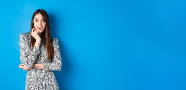 Coquettish girl in trendy long dress looking excited at camera, standing on blue background