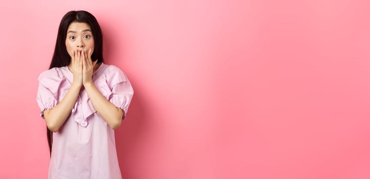 Shocked asian girl with long dark hair, gasping and covering mouth with hands, look at bad news, terrible accident, standing on pink background