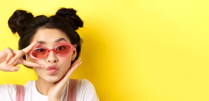 Summer fashion concept. Cute and glamour asian girl in sunglasses, showing v-sign kawaii and looking at camera, standing on yellow background