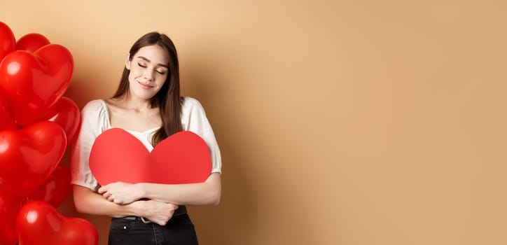 Romantic woman hugging big red heart and smiling dreamy, falling in love on Valentines day, dreaming about lover, standing on beige background near balloons.