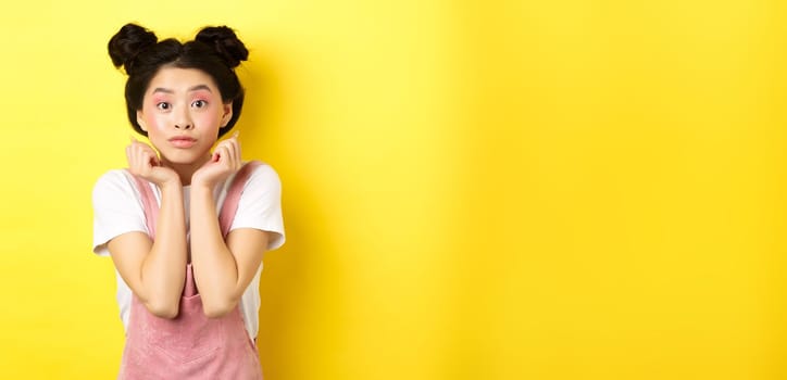 Cute teenage asian girl with beauty makeup, listen with interest, lean on hands and looking excited at camera, standing on yellow background