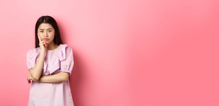 Bored asian teen girl look indifferent at camera, lean face on hand in skeptical pose, standing reluctant against pink background