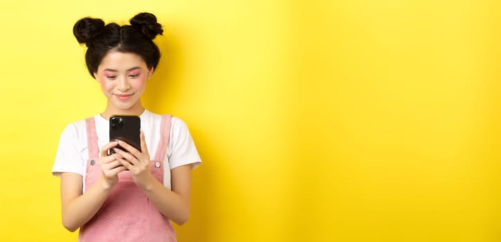 Stylish asian girl with glamour makeup using mobile phone, smiling at screen, standing on yellow background