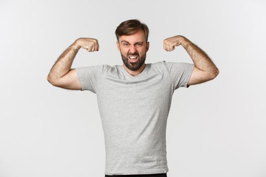 Portrait of confident bearded man flexing biceps, showing his muscles after gym workout, standing over white background