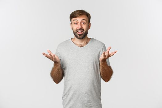 Portrait of handsome bearded male moden in grey t-shirt, raising hands up and looking amazed, congratulating you, standing over white background