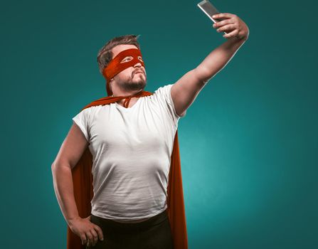 Super Hero man Takes Selfie By Mobile Phones. Man In A Super Hero Costume In A Red Mask And Cloak Isolated On Biscay Green Background