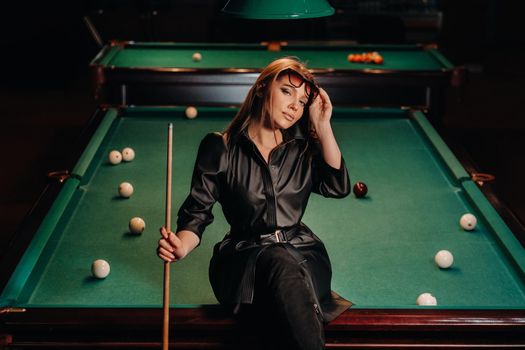 A girl with glasses sits on a pool table in a club.Russian billiards