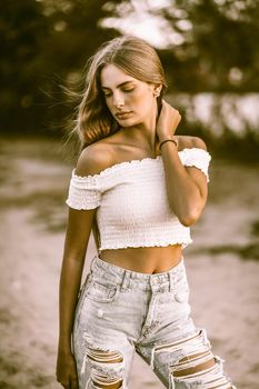 Fashion teenage girl wearing jeans and white top posing in evening on nature background. Vogue concept. Toned image