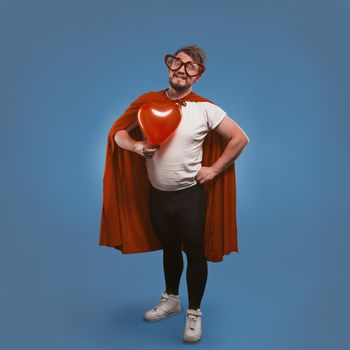 Romantic super hero holds big heart. Happy man in love wearing superhero costume and heart-shaped glasses isolated on blue background. Valentine's day concept. Copy spase on both sides