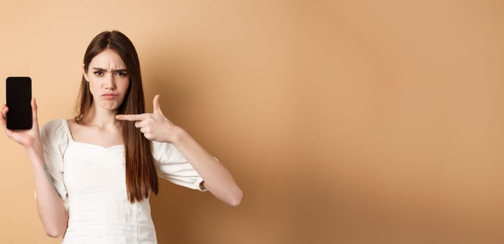Disappointed frowning girl pointing at empty phone screen, complaining at online news, standing on beige background
