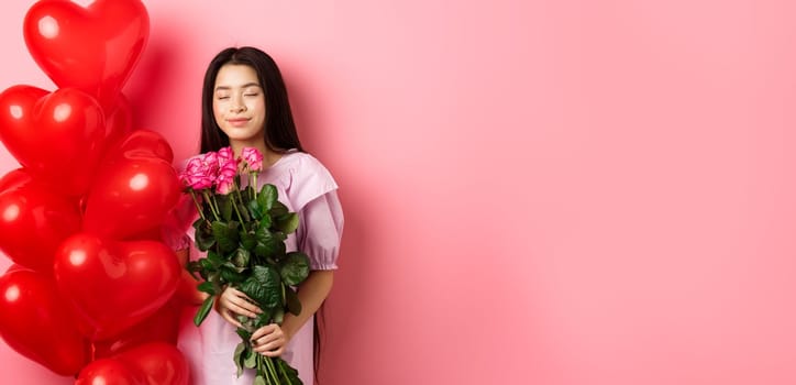 Valentines day concept. Romantic teen asian girl dreaming of love or date, close eyes and smile, holding flowers from lover, receive bouquet of roses and red heart balloons, pink background.