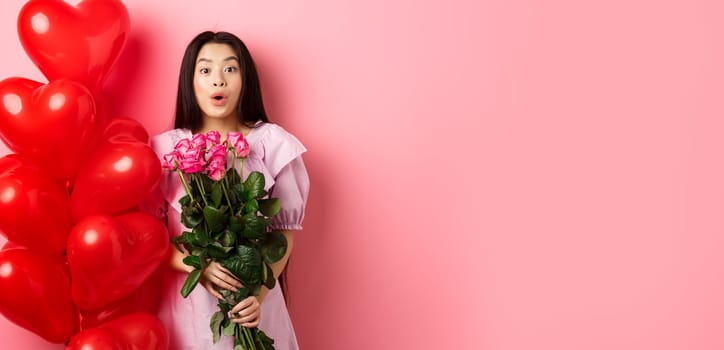 Surprised asian girl in dress standing near valentines day heart balloons and say wow at camera, holding flowers bouquet from lover, romantic date with roses, pink background