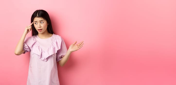 Are you stupid. Shocked asian woman pointing at head and complaining, scolding someone crazy or strange, look aside, standing against pink background