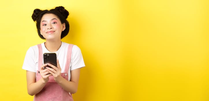 Cute stylish asian girl using mobile phone, wearing glamour pink makeup and summer clothes, standing on yellow background