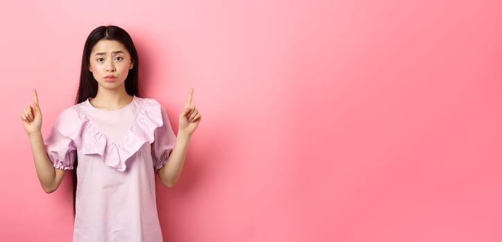 Cute timid asian woman pointing fingers up, frowning and look upset, pointing fingers up at logo, standing in dress on pink background