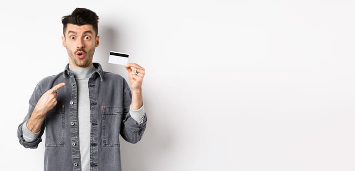 Wow look here. Excited guy pointing at plastic credit card and look amazed with awesome deal, standing against white background