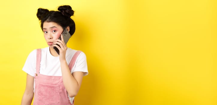 Cute teen girl talking on smartphone, making silly pouting face and look timid at camera, standing with glamour makeup on yellow background