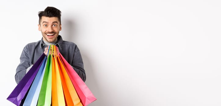 Excited smiling guy holding colorful shopping bags and rejoicing with discounts in store, standing against white background