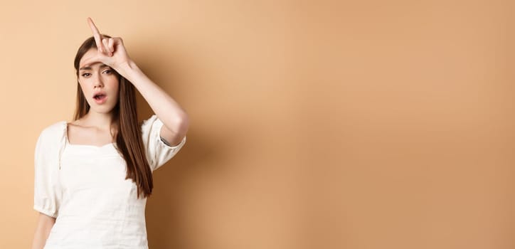 Confident woman mocking people with loser sign, being mean, standing on beige background