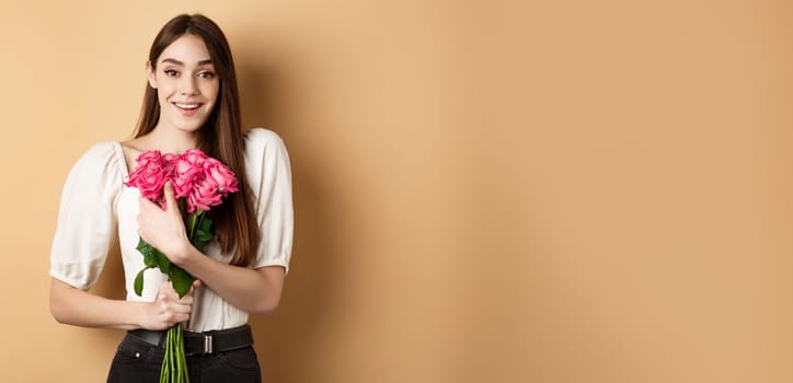 Valentines day. Romantic girl smiling happy at camera, holding bouquet of pink roses from lover, standing on beige background.
