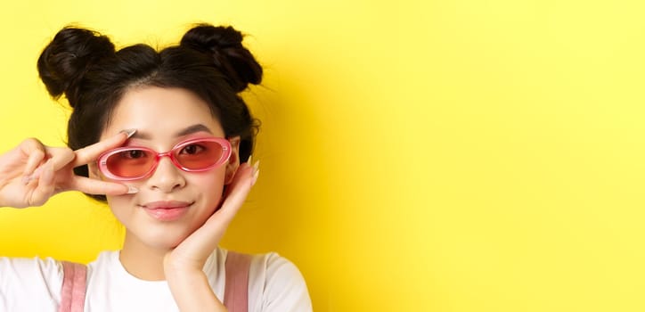 Summer fashion concept. Close-up of trendy korean girl with hairbuns and sunglasses, showing v-sign on vacation, standing against yellow background.