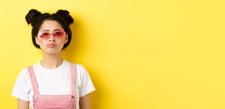 Summer and fashion concept. Bored asian teen girl in sunglasses pouting, standing moody on yellow background