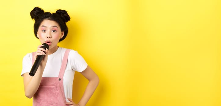 Cute asian teen girl with bright makeup, singing in microphone karaoke, standing against yellow background