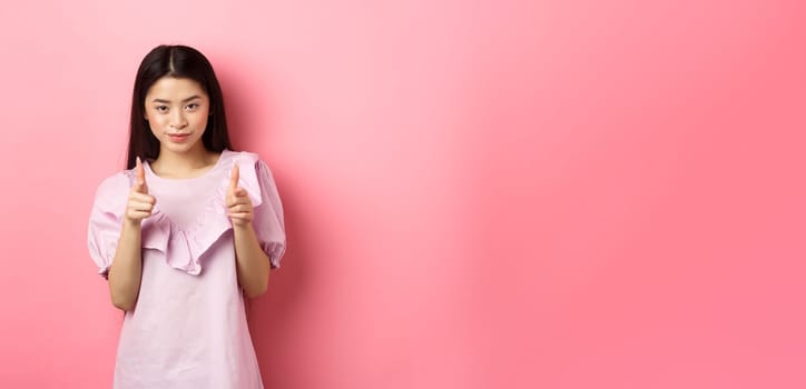 Sassy asian woman pointing at camera, smiling and inviting you, beckon or praise person, standing on pink background
