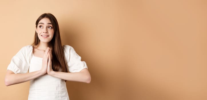 Hopeful young girl begging god and looking dreamy at upper left corner, praying or making wish, standing on beige background