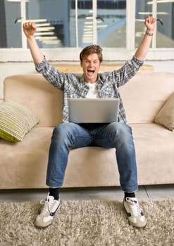 Oh yeah Ive got something to celebrate. A young man with his arms raised in celebration while using his laptop.