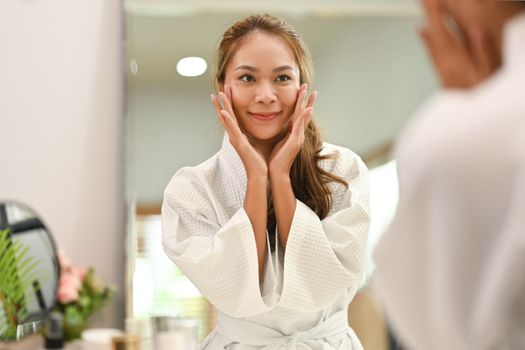 Pleased young woman wearing white bathrobe applying softening moisturizing face cream for skin face care in front of mirror
