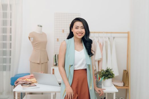 Young Asian fashion designer smiling and standing in front of the desk in the workshop
