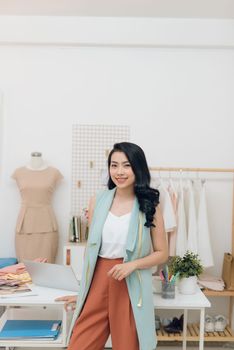 Young Asian fashion designer smiling and standing in front of the desk in the workshop