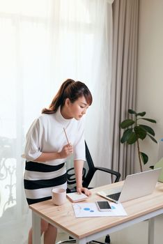 Young Asian woman standing to write in notebook while looking at laptop screen at home.