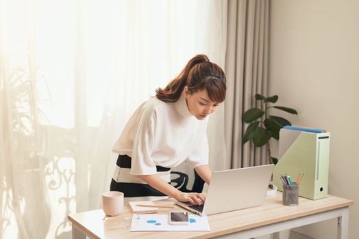 Young Asian businesswoman working with laptop and standing behind the desk at home.