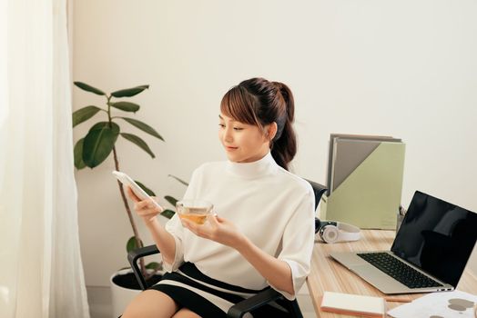 Young Asian woman using phone at her office while drinking tea 