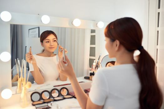 Young Asian woman applying make up (paint her eyelashes) in front of a mirror. Focus on her reflection
