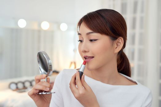  Smiling young Asian woman applying lipstick and looking to mirror.