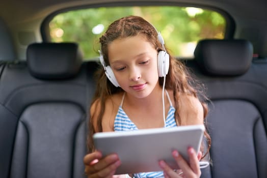 Keeping the car trip an entertained one. a young girl sitting in a car wearing headphones and using a digital tablet.