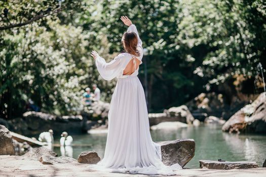 a beautiful woman in a long white dress looks into the distance at a beautiful lake with swans rear view