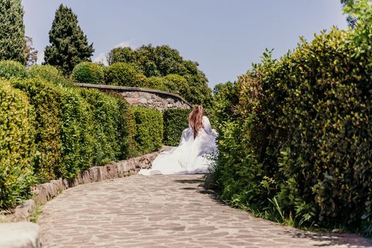 Brunette runs white dress park. A beautiful woman with long brown hair and a long white dress runs along the path along the beautiful bushes in the park, rear view