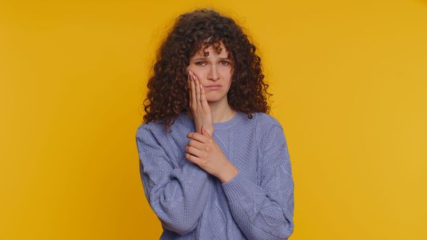 Hipster curly haired woman touching sore cheek suffering from toothache cavities or gingivitis waiting for dentist appointment gums disease. Young girl indoor studio shot isolated on yellow background