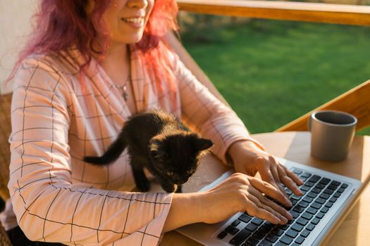 Work from home or study online and videocall with a woman gardener working at laptop outdoor on terrace with kitten close-up, sitting on ratang chair around houseplant and flower pots