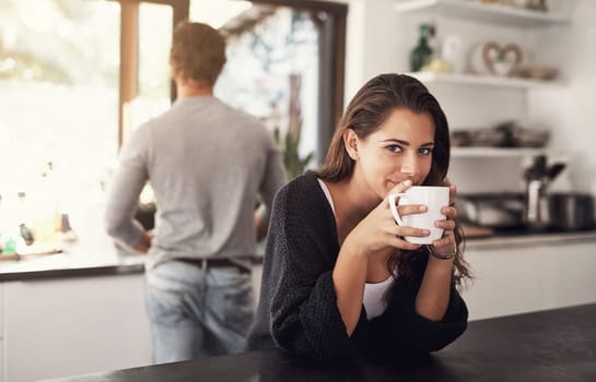 Coffee puts the mmm in mornings. Portrait of a young woman having coffee in the kitchen with her husband standing in the background.