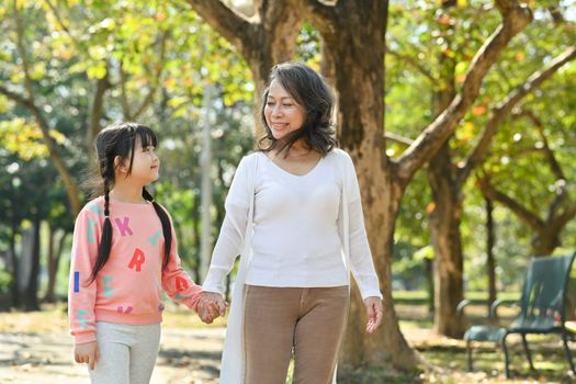 Smiling middle age grandmother and cute granddaughter walking in public park at morning