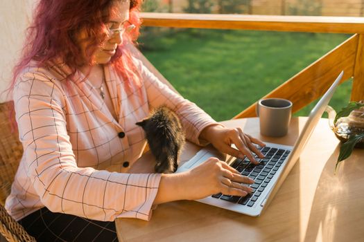 Work from home or study online and videocall with a woman gardener working at laptop outdoor on terrace with kitten, sitting on ratang chair around houseplant and flower pots