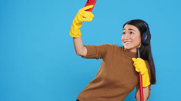 Cheerful housekeeper holding cleaning broom while taking selfie with smartphone