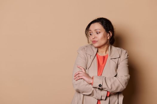 Unsure woman with folded hands looks away, apprehensive, expressing insecurity and worry over beige background. Ad space