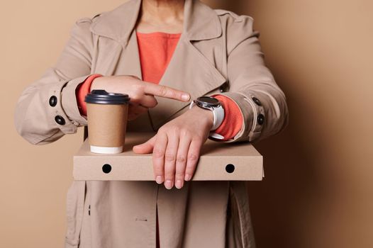 It's lunch time. Close-up woman holding takeaway pizza and coffee in paper mug, points her index finger at a wristwatch