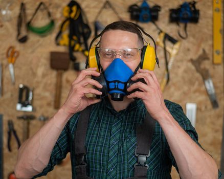 A carpenter in goggles and earmuffs removes a protective respirator.
